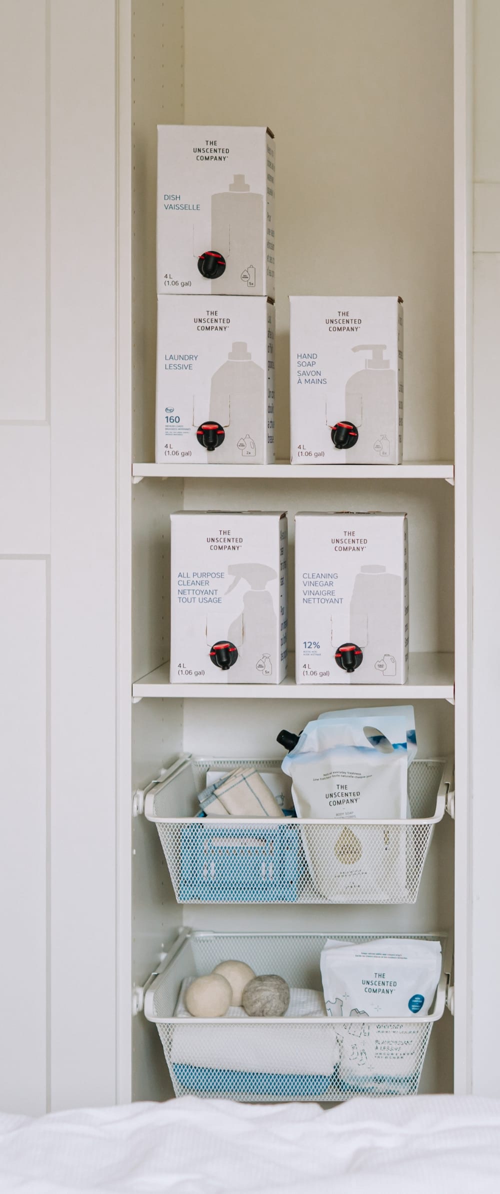 A bright, organized laundry room with a white shelving unit storing The Unscented Company refill boxes — including dish soap, laundry detergent, hand soap, and all-purpose cleaner — alongside wire baskets holding smaller products and dryer balls, next to a fiddle-leaf fig plant.