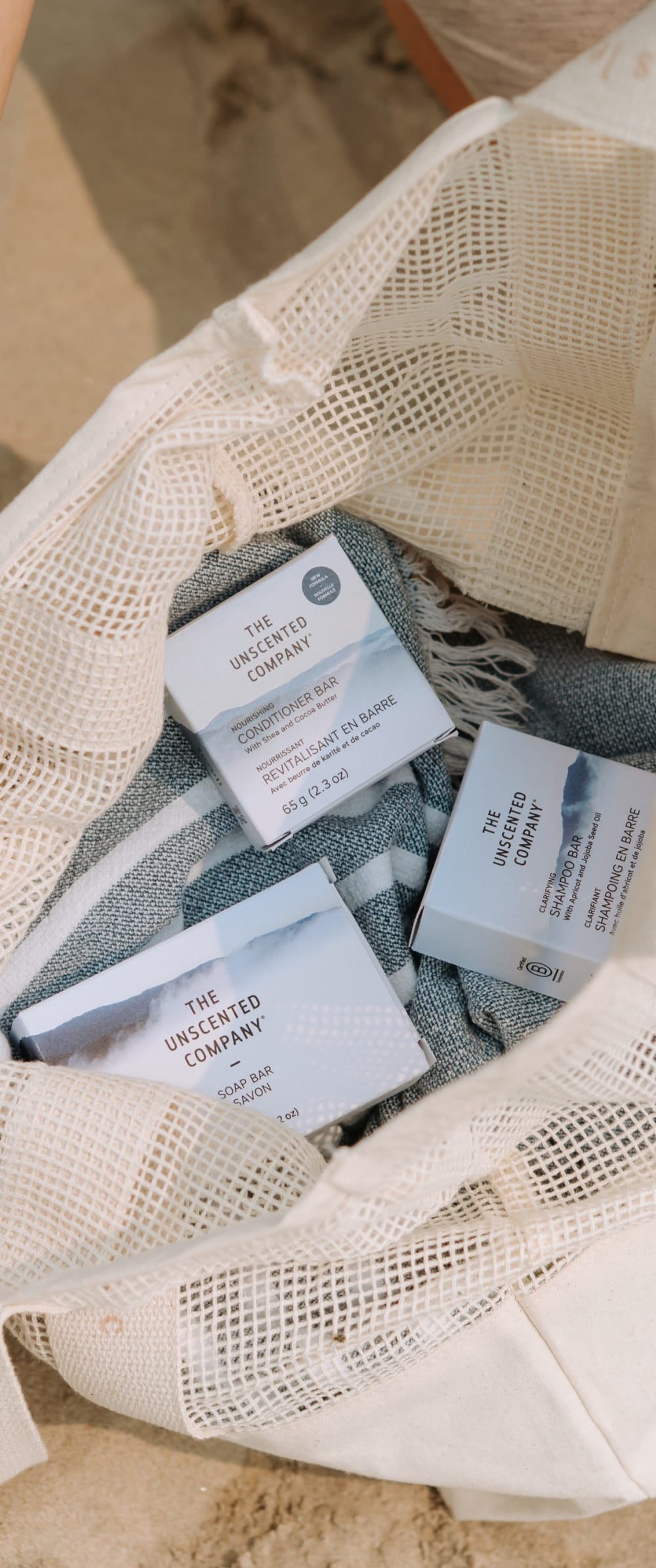 Un sac en filet de coton sur le sable contenant les soins zéro déchet de The Unscented Company : shampoing en barre, revitalisant en barre et savon.