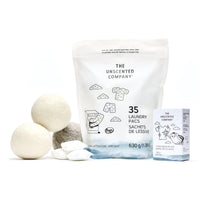 Une composition épurée sur fond blanc présentant un sac de 35 sachets de lessive (630 g), une boîte de barre détachante (140 g) et trois balles de séchage en laine (deux blanches, une grise) avec quelques sachets de lessive individuels au premier plan.