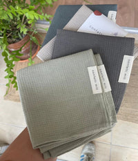 A handful of waffle-weave dish towels in various neutral shades (sage green, beige, dark grey, light grey, and navy), each labeled "confetti mill," held by hand over a tiled floor, with a potted green plant visible in the background.