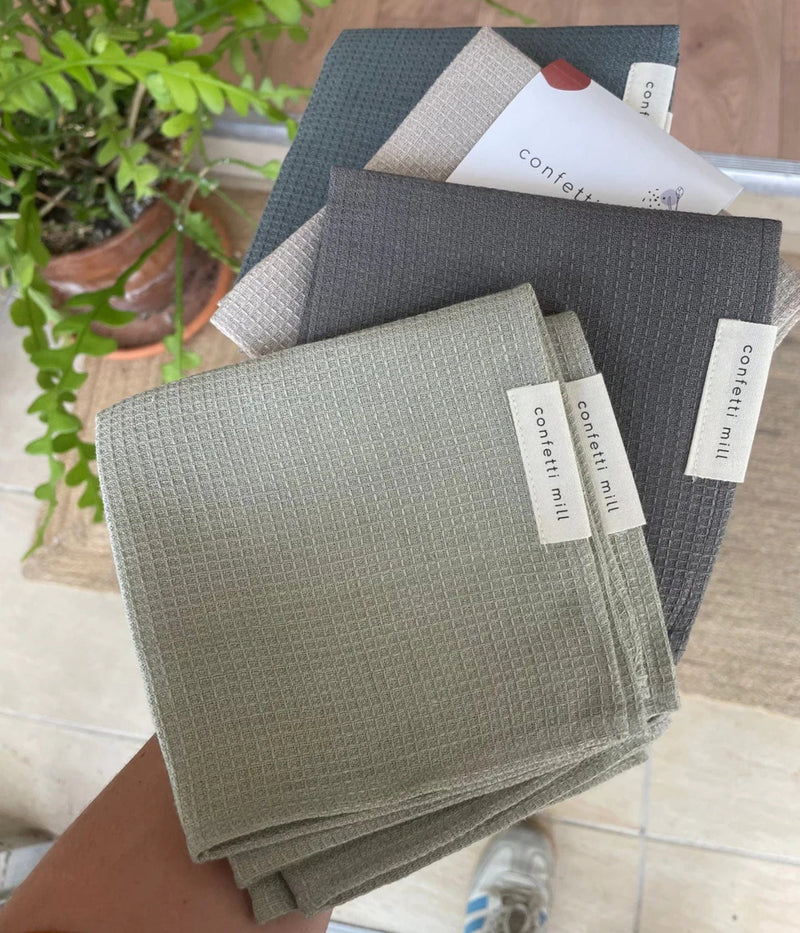 A handful of waffle-weave dish towels in various neutral shades (sage green, beige, dark grey, light grey, and navy), each labeled "confetti mill," held by hand over a tiled floor, with a potted green plant visible in the background.
