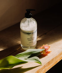 A bottle of The Unscented Company hand soap filled with bubbles, placed on a sunlit wooden surface beside a single pink tulip with green leaves.