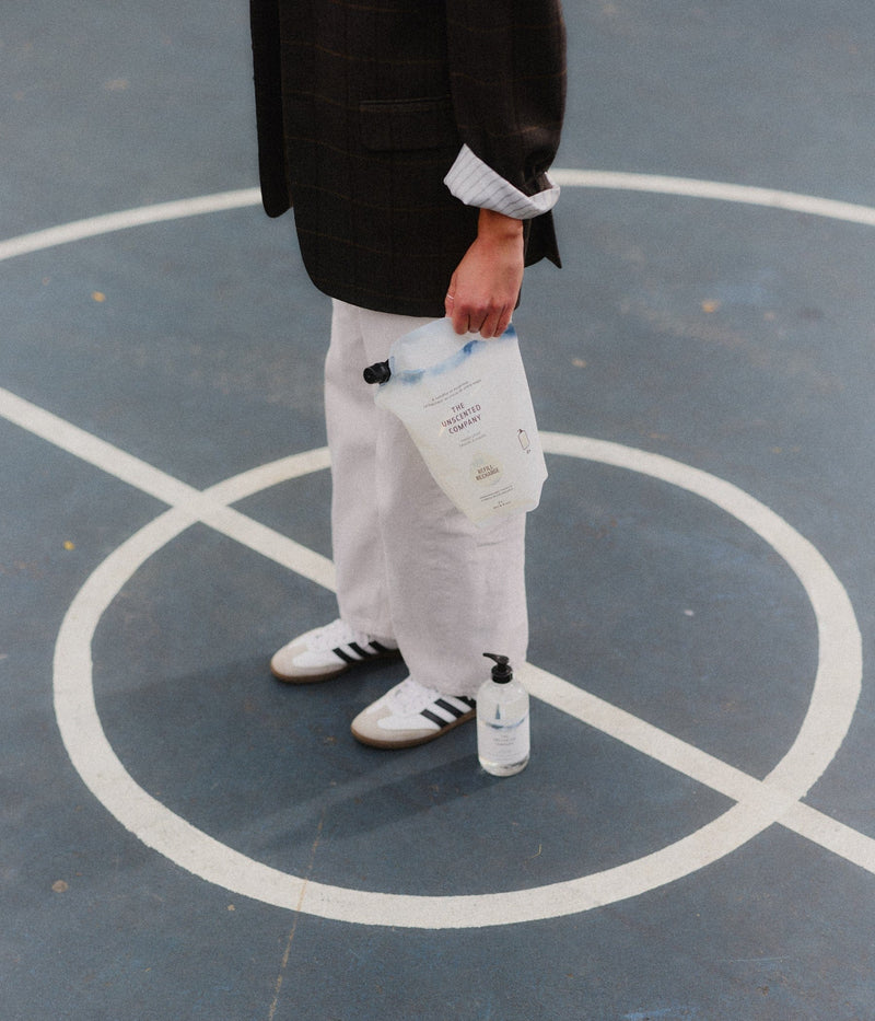 Person standing at the center of a basketball court, holding a refill pouch of The Unscented Company hand soap. A bottle of hand soap is placed on the ground by their feet.