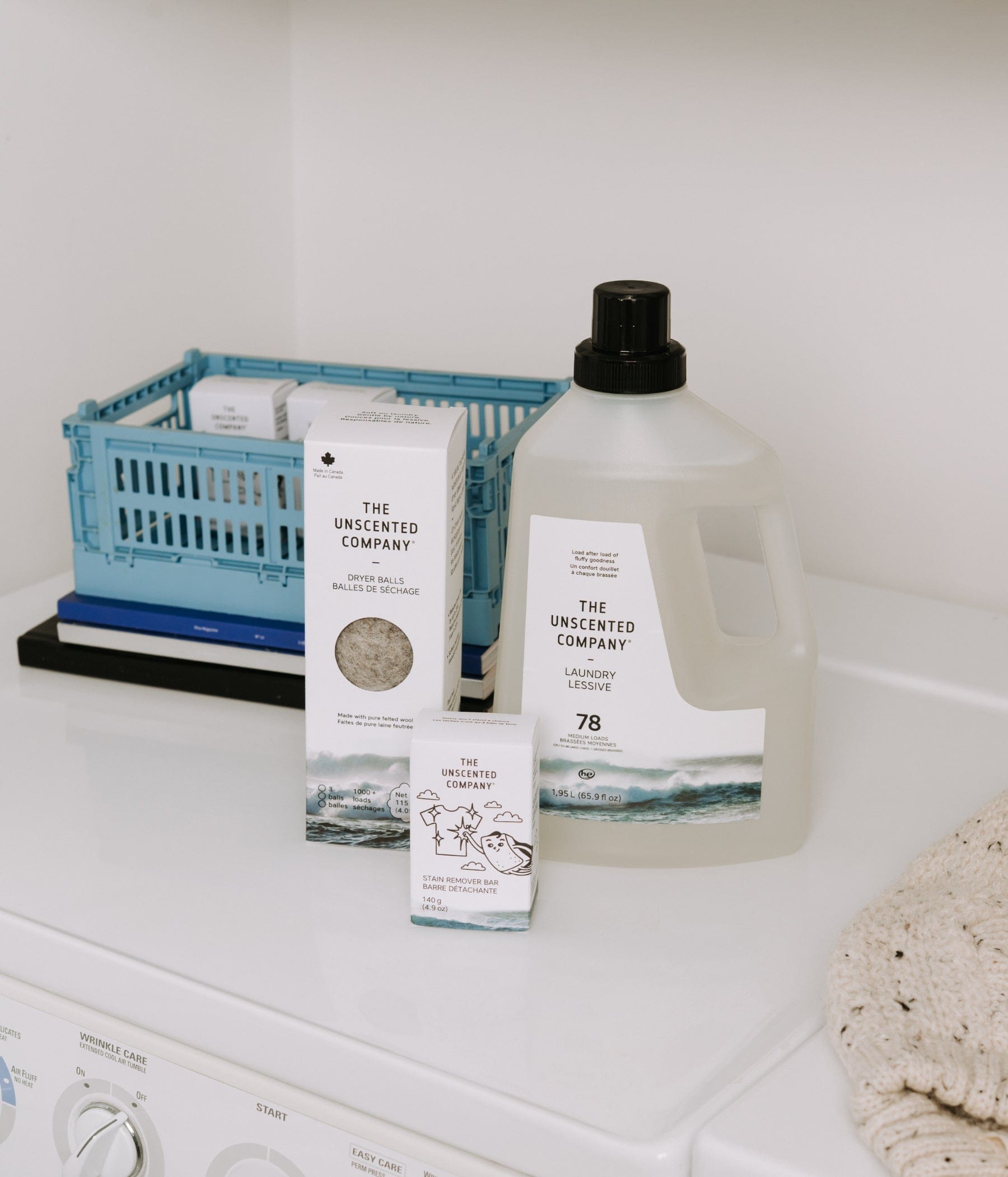 Un assortiment de produits The Unscented Company comprenant un sac de 35 sachets de lessive, une boîte de balles de séchage et une barre détachante, disposé sur une machine à laver blanche à côté d'un chandail en tricot. En arrière-plan, une étagère contient des serviettes blanches et une boîte de recharge de 4 L.