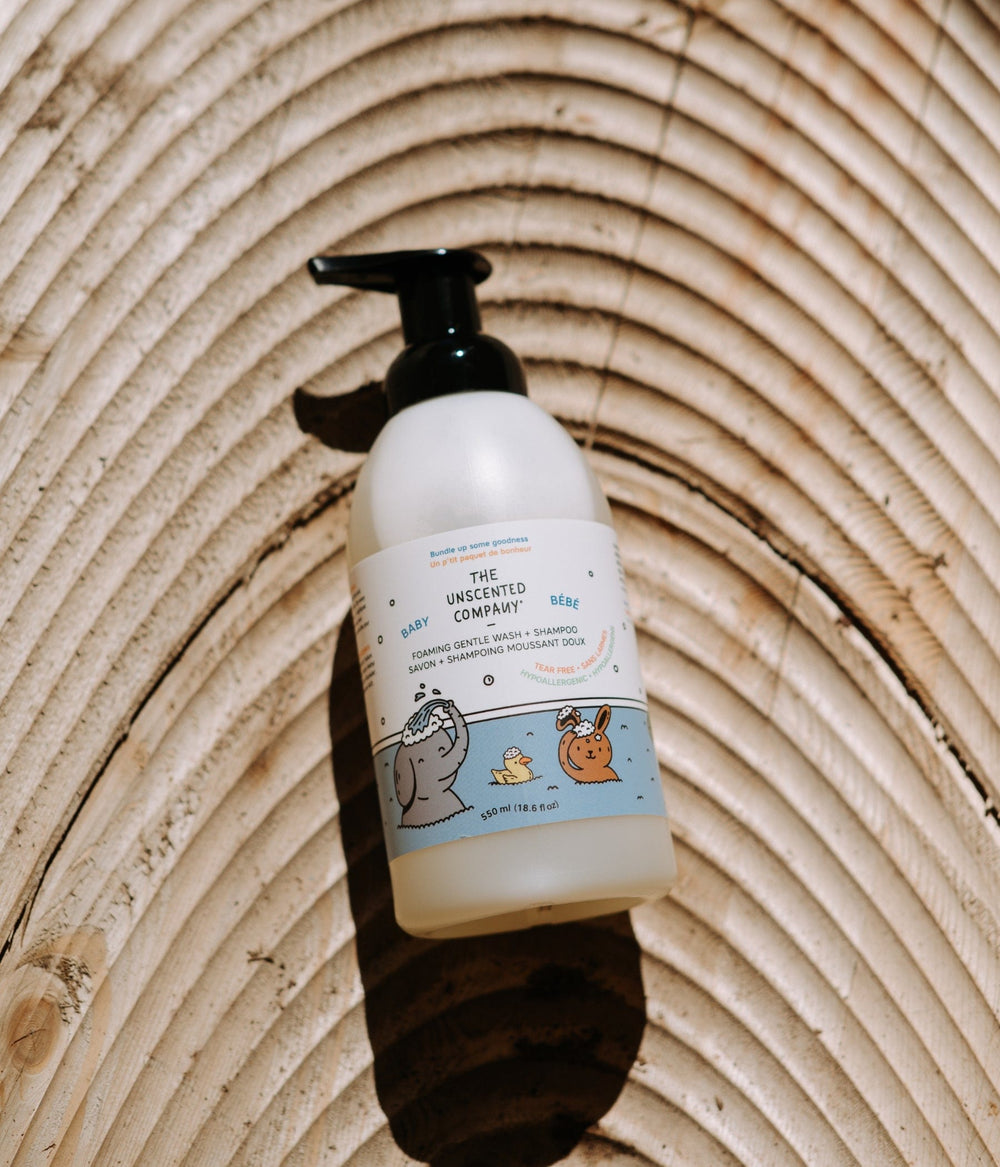 A pump bottle of The Unscented Company Foaming Gentle Wash + Shampoo for Baby (550 ml) resting on a textured wooden surface with concentric ring patterns. The label features playful cartoon illustrations of an elephant, a duck, and a rabbit in a bath.