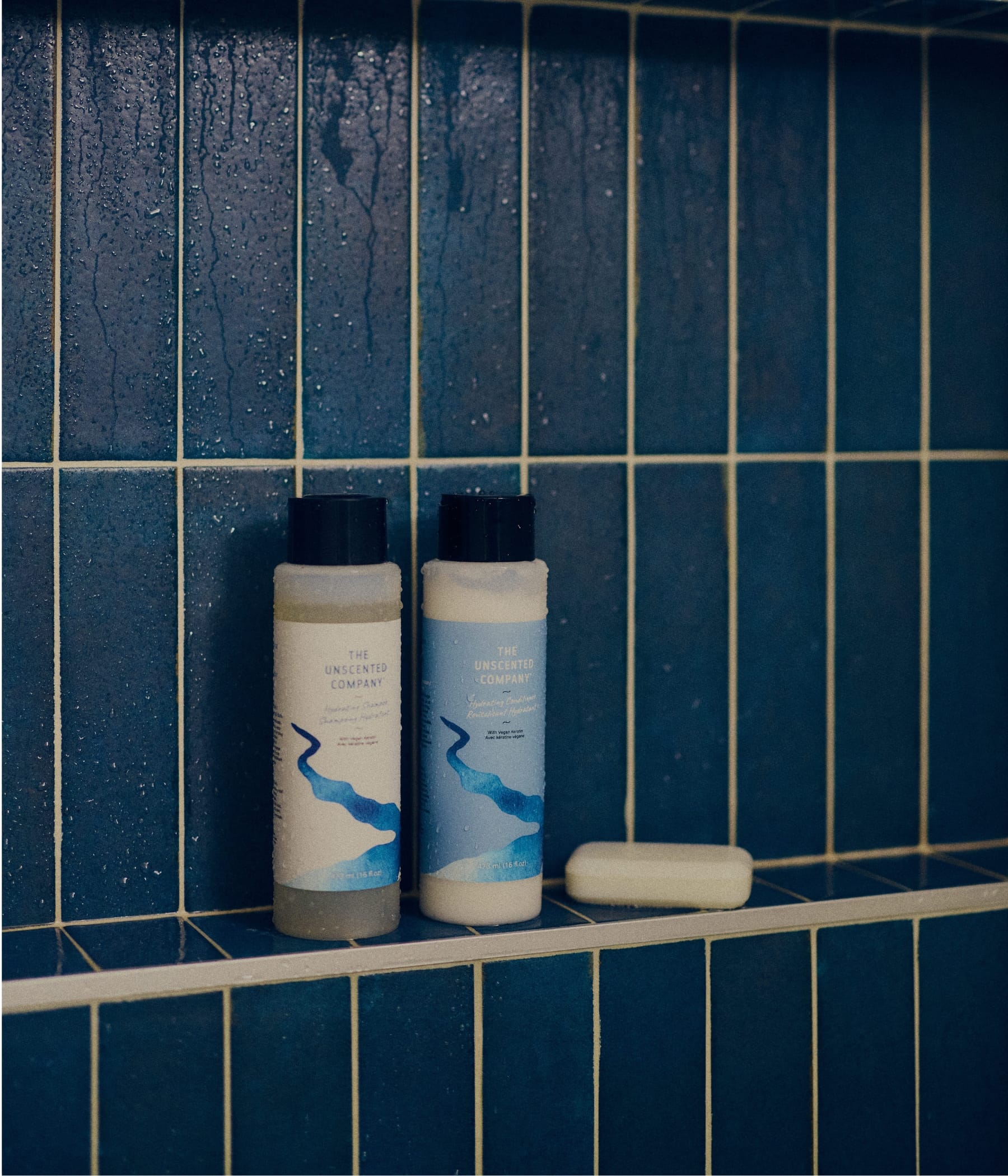 Les bouteilles de shampoing hydratant et de revitalisant hydratant de The Unscented Company sur une tablette de douche mouillée, avec un savon en barre, devant un carrelage vertical bleu avec des gouttes d'eau.