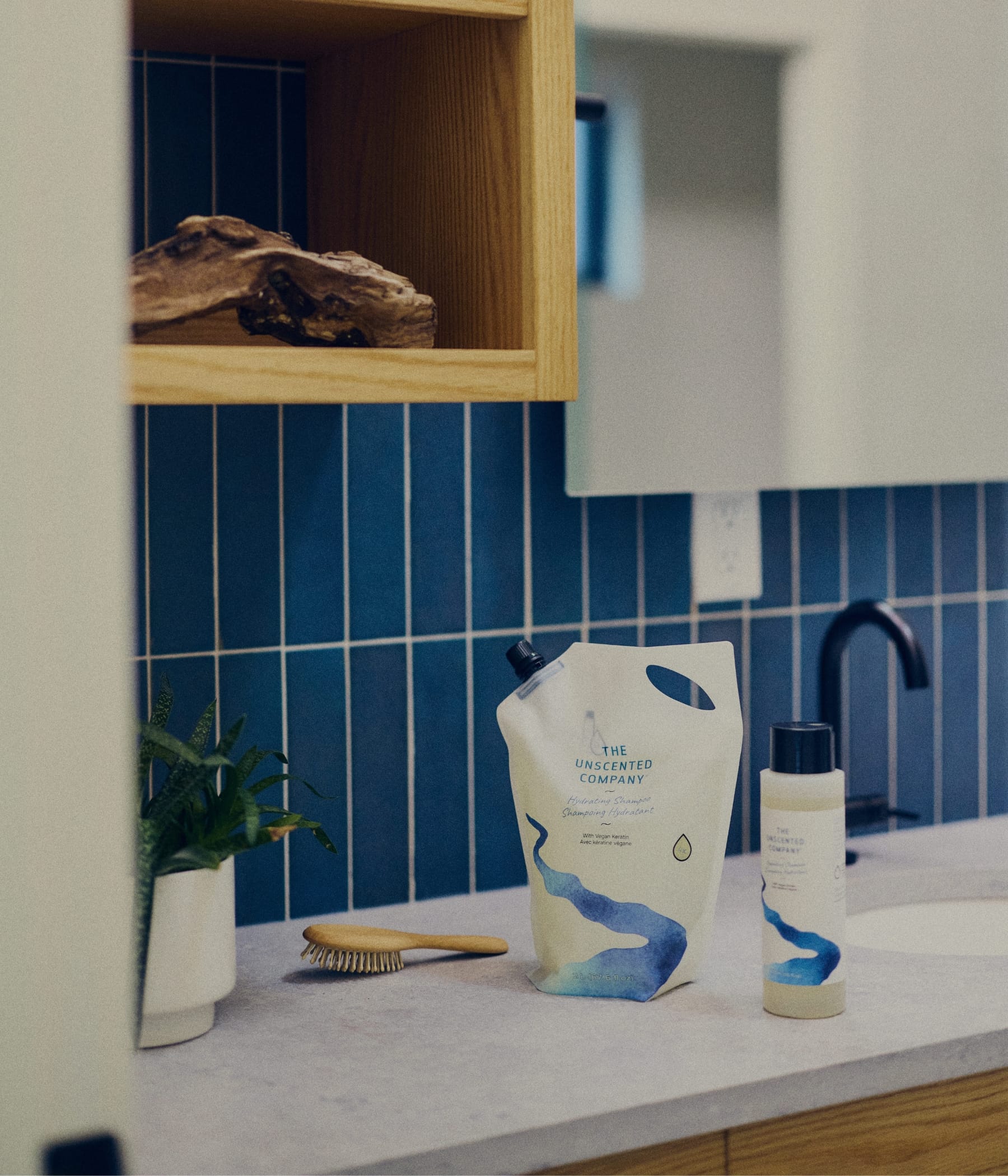 The Unscented Company Hydrating Shampoo refill pouch and travel-size bottle on a bathroom counter, alongside a wooden hairbrush and a small plant, with blue vertical tile and a wood shelf in the background.