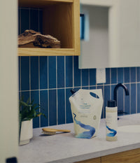The Unscented Company Hydrating Shampoo refill pouch and travel-size bottle on a bathroom counter, alongside a wooden hairbrush and a small plant, with blue vertical tile and a wood shelf in the background.