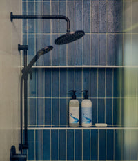 The Unscented Company Nourishing Shampoo and Nourishing Conditioner pump bottles on a shower shelf alongside a bar of soap, with a matte black rain showerhead and blue vertical tile in the background.