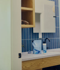 A refill pouch and bottle of The Unscented Company hair care products sit on a white bathroom countertop beside a blue hair clip. The minimalist white-and-blue packaging is displayed in front of a sink with a matte black faucet, blue tiled wall, mirror, and natural wood cabinetry.