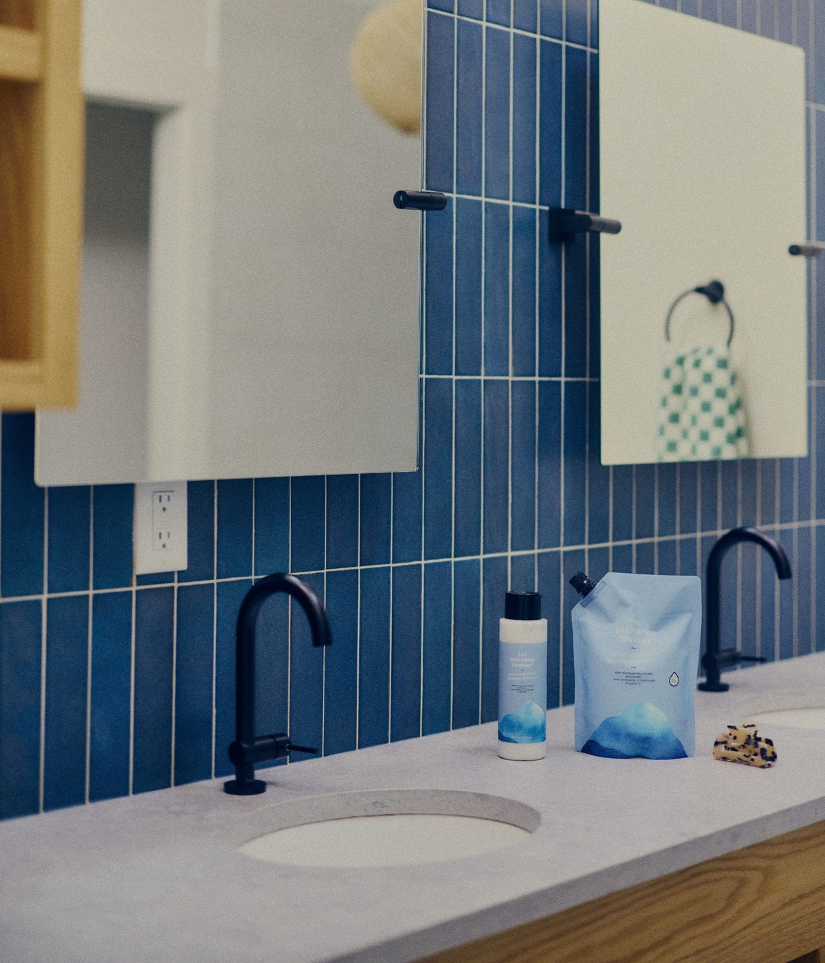 A modern bathroom countertop with two white sinks, matte black faucets, rectangular mirrors, and a blue tiled wall. Between the sinks are a 473 mL bottle and 1-litre refill pouch of The Unscented Company volumizing conditioner with blue graphics, formulated with hydrolyzed rice protein and glycerin, alongside a small patterned hair clip.