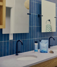 A modern bathroom countertop with two white sinks, matte black faucets, rectangular mirrors, and a blue tiled wall. Between the sinks are a 473 mL bottle and 1-litre refill pouch of The Unscented Company volumizing conditioner with blue graphics, formulated with hydrolyzed rice protein and glycerin, alongside a small patterned hair clip.