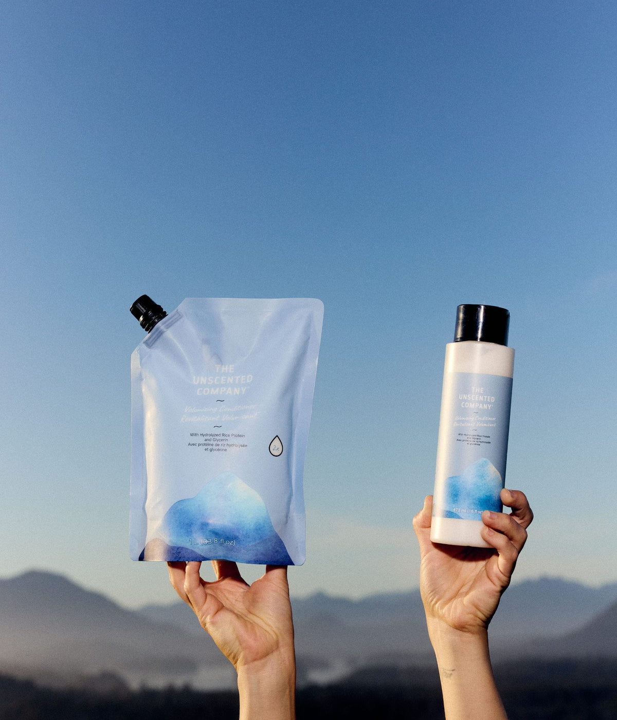 Two hands hold The Unscented Company volumizing conditioner products against a clear blue sky, including a 1-litre refill pouch and a 473 mL bottle with blue botanical graphics. The unscented hair conditioner is formulated with hydrolyzed rice protein and glycerin, with a hazy mountain range visible in the background.