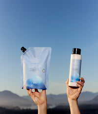 Two hands hold The Unscented Company volumizing conditioner products against a clear blue sky, including a 1-litre refill pouch and a 473 mL bottle with blue botanical graphics. The unscented hair conditioner is formulated with hydrolyzed rice protein and glycerin, with a hazy mountain range visible in the background.