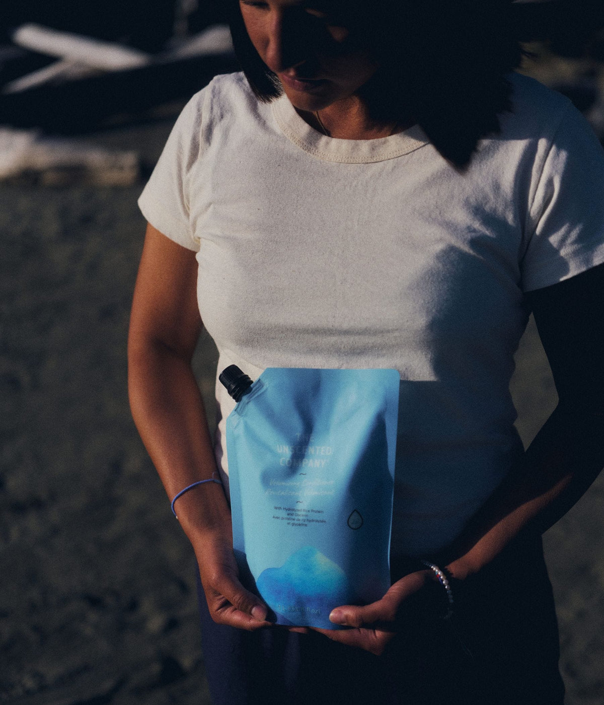 A woman with short dark hair wearing a white T-shirt holds a 1-litre refill pouch of The Unscented Company volumizing conditioner in front of her torso outdoors. The blue hair care refill pouch features botanical graphics and is formulated with hydrolyzed rice protein and glycerin, illuminated by warm sunlight and deep shadows.