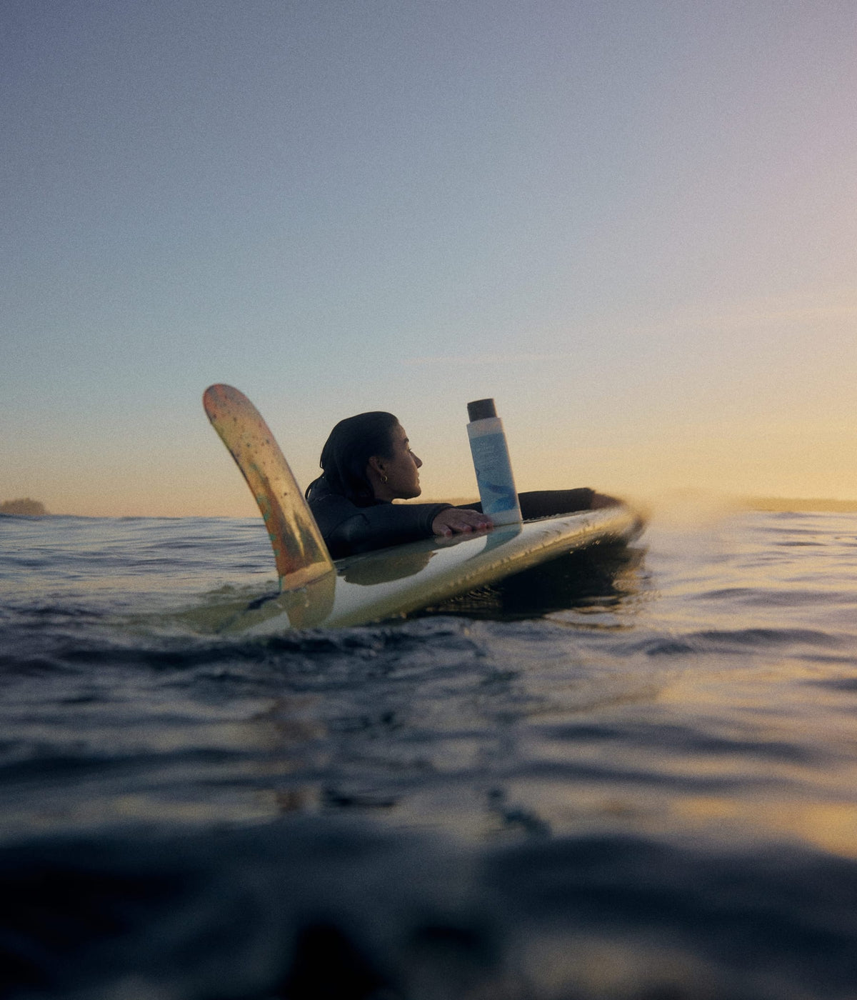A surfer resting on her surfboard in the water at sunset, next to a bottle of hydrating shampoo from The Unscented Company.