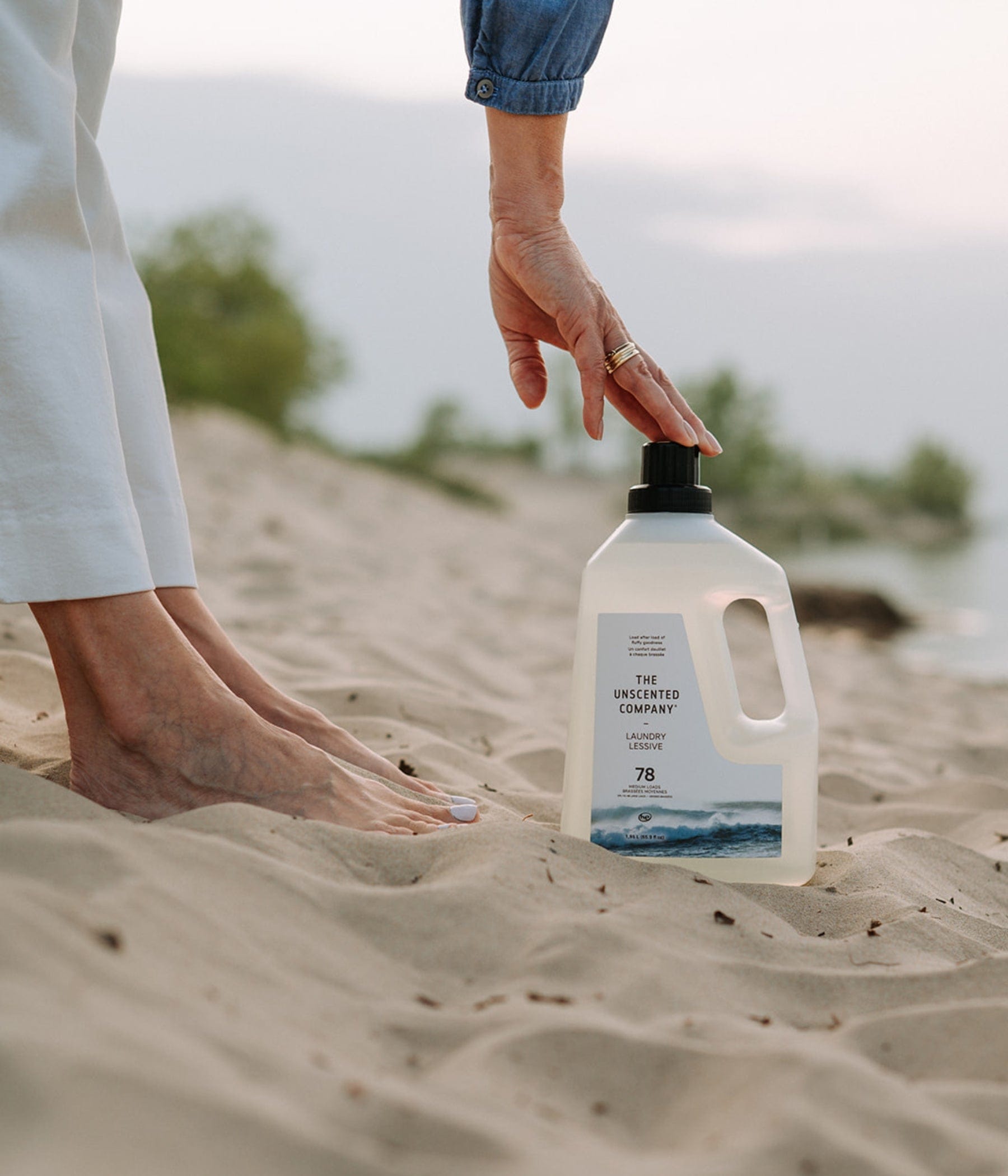 A woman's hand touching the cap of a bottle of The Unscented Company laundry detergent, placed on the sand near her bare feet.