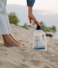 A woman's hand touching the cap of a bottle of The Unscented Company laundry detergent, placed on the sand near her bare feet.