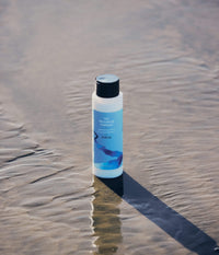 A bottle of The Unscented Company hydrating conditioner standing on wet beach sand.