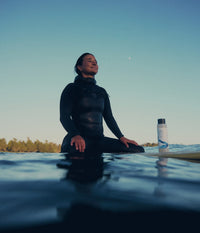 Une surfeuse assise sur sa planche dans l'eau au coucher du soleil, à côté d'une bouteille de shampoing hydratant The Unscented Company.