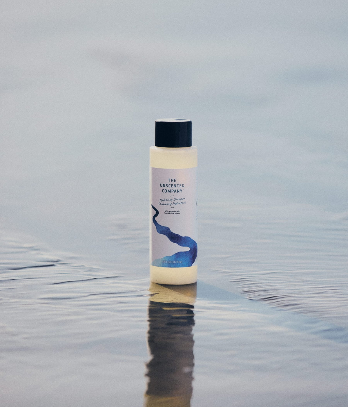 Une bouteille de shampoing hydratant The Unscented Company posée debout sur le sable mouillé d'une plage.