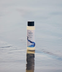 Une bouteille de shampoing hydratant The Unscented Company posée debout sur le sable mouillé d'une plage.
