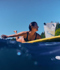 A woman laying on a surfboard in the water next to the hydrating shampoo refill pouch from The Unscented Company under a bright blue sky.