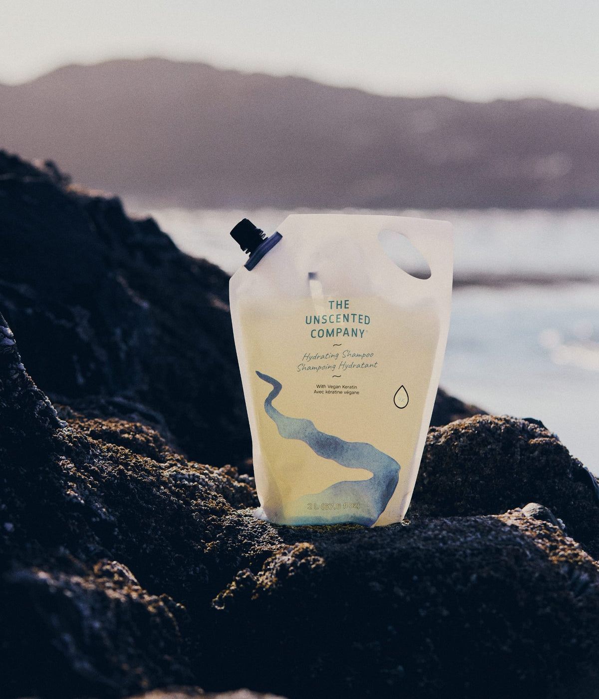 A 2L refill pouch of The Unscented Company Hydrating Shampoo resting on dark coastal rocks by the sea.