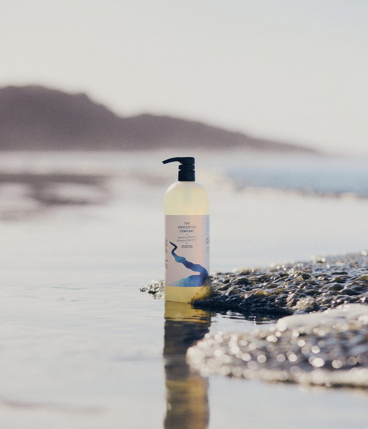 Une bouteille à pompe 946ml du shampoing hydratant The Unscented Company posée dans l'eau au bord d'une plage rocheuse.