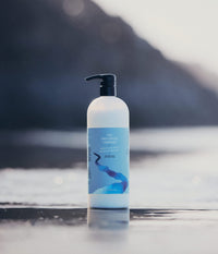 A 946ml pump bottle of The Unscented Company Hydrating Conditioner standing on wet beach sand near the shoreline.