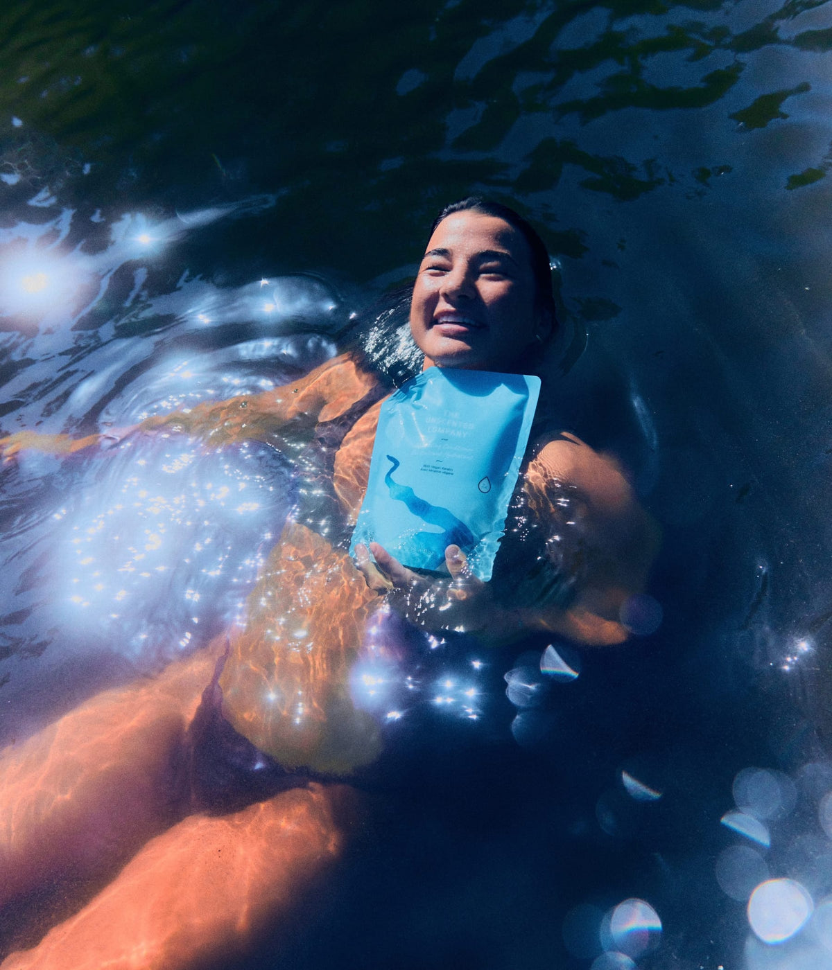 A smiling woman floating on her back in shimmering water holding a 1L refill pouch of The Unscented Company hydrating conditioner.