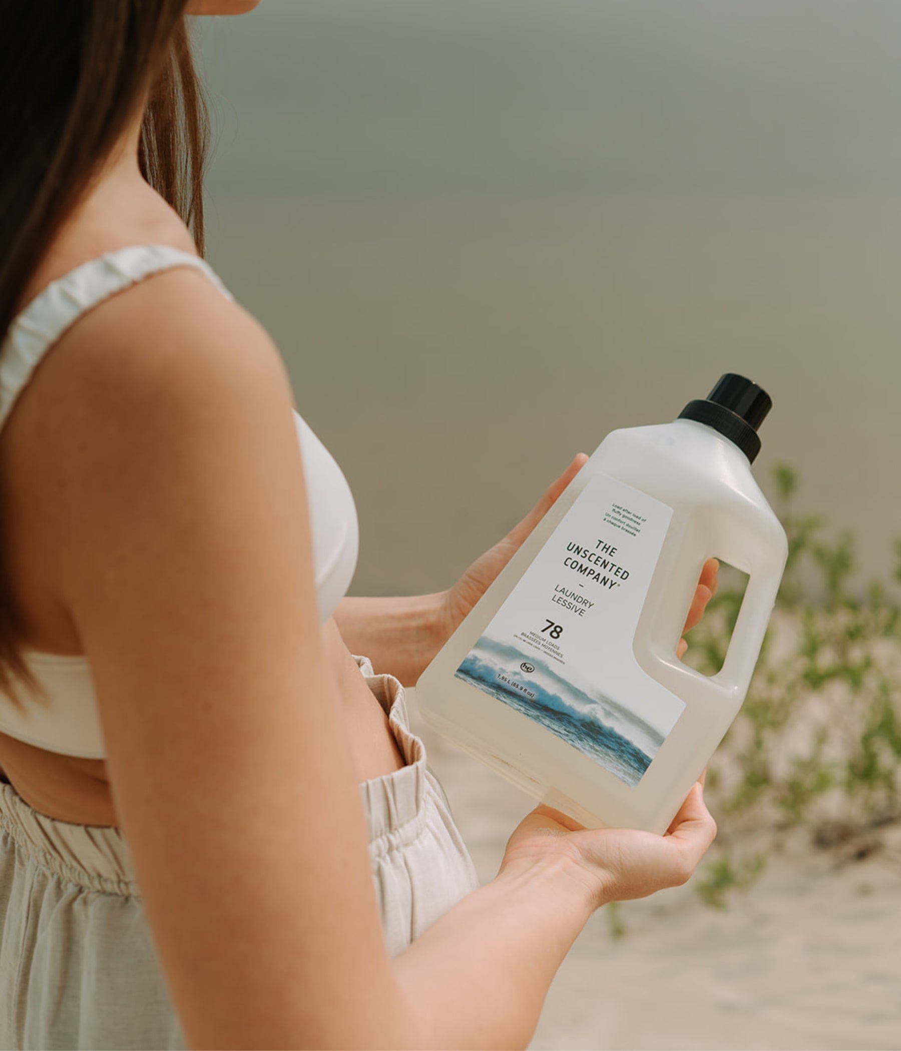 Woman holding a bottle of The Unscented Company laundry detergent on a sandy beach, near a calm lake.