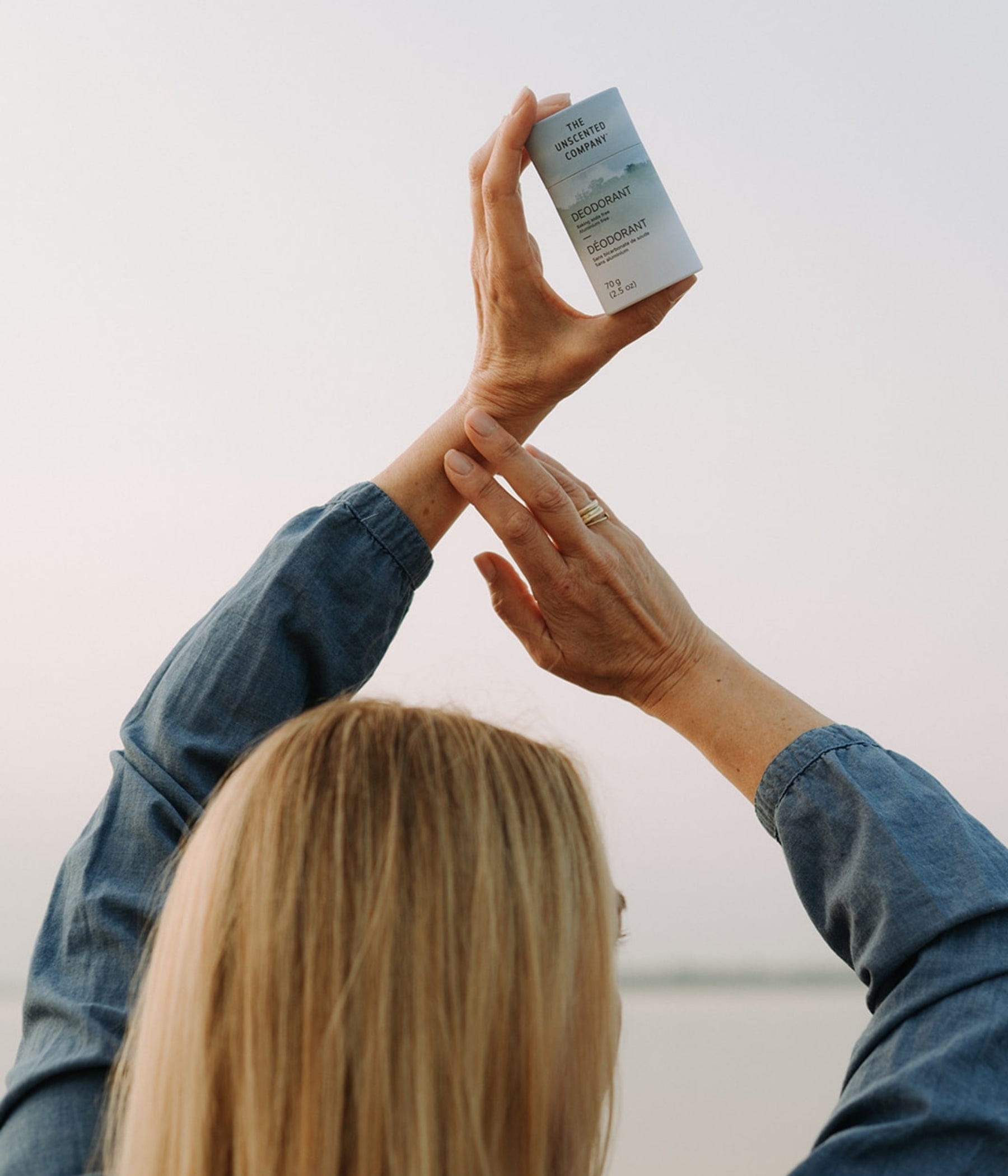 Woman seen from behind holding up The Unscented Company deodorant to the sky at the beach.