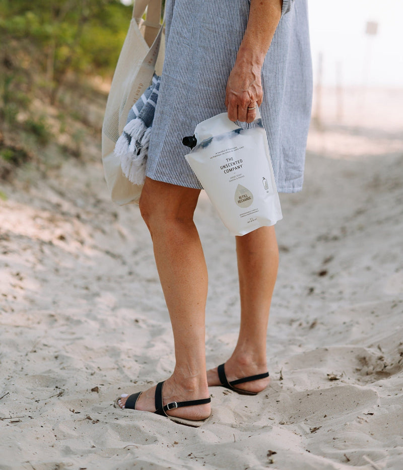 A person walking on a sandy path, holding a refill pouch of The Unscented Company hand soap and a tote bag with a towel.
