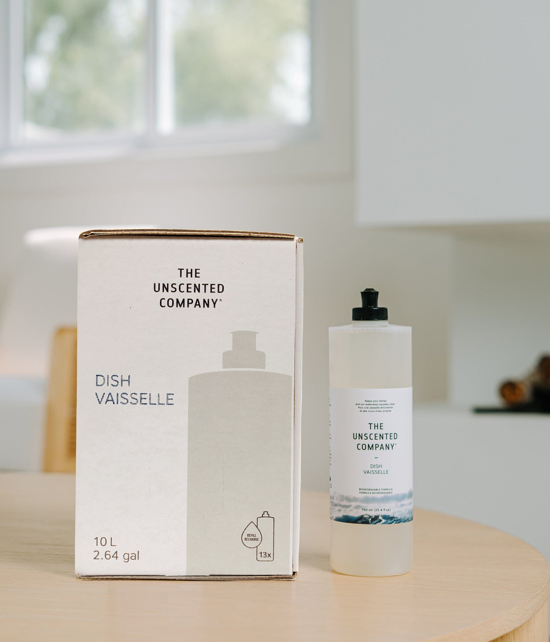 Refill box and bottle of The Unscented Company Dish / Vaisselle dish soap on a wooden table, showing 10 L (2.64 gal) refill packaging with 13x refill icon and 750 ml bottle, photographed in a bright minimalist kitchen.