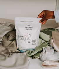 Gros plan d'une main tenant un sac de lessive pour vêtements délicats The Unscented Company, déposé sur un amas de tissus soyeux et satinés aux tons vert sauge, gris et crème.