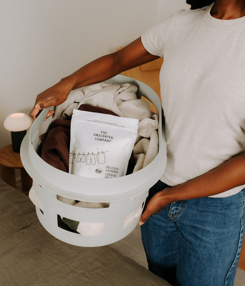 A person wearing a white t-shirt and jeans holds a grey laundry basket filled with clothes and a bag of The Unscented Company delicate laundry detergent.