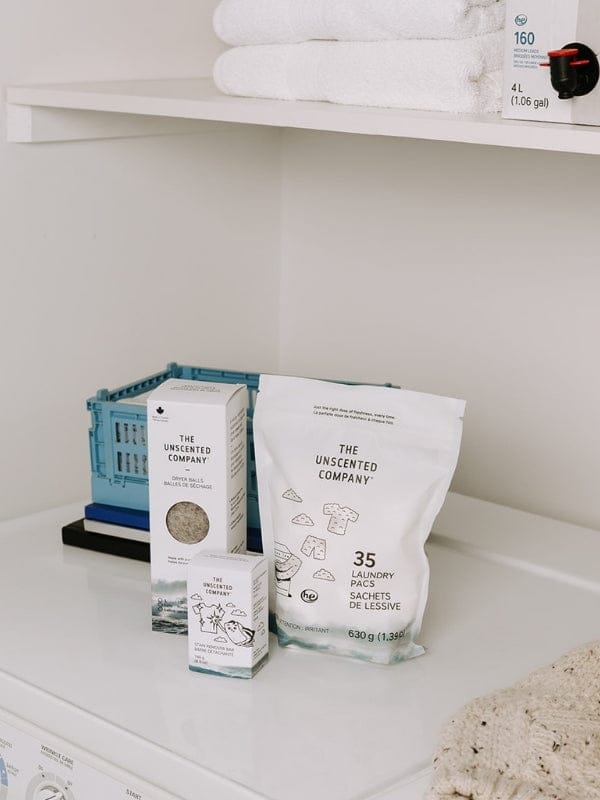 Un ensemble de produits The Unscented Company (balles de séchage, sachets de lessive et barre détachante) rangés sur une machine à laver blanche dans une buanderie épurée.
