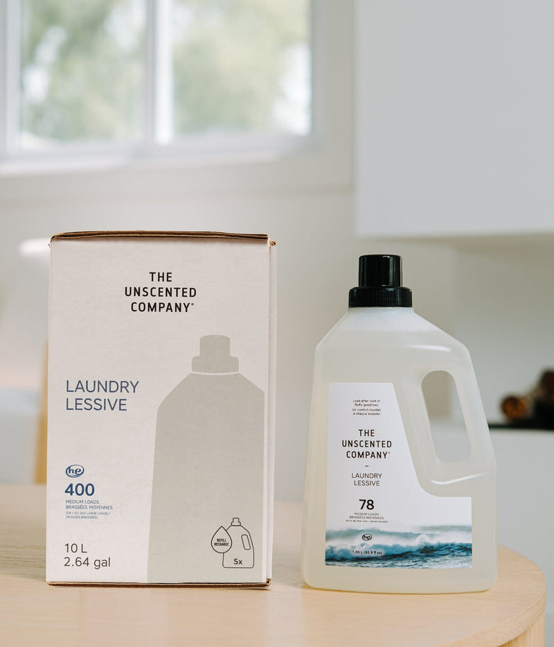 Boîte recharge 10 L (2,64 gal) et bouteille de lessive The Unscented Company posées sur une table en bois dans une cuisine lumineuse, avec design blanc minimaliste, texte « LAUNDRY / LESSIVE », icône 400 brassées moyennes et symbole recharge 5x.