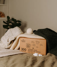 Une grande boîte de 300 sachets de lessive The Unscented Company entourée de chandails en laine, d'une balle de séchage et de trois sachets individuels posés sur un tissu texturé dans un décor chaleureux.