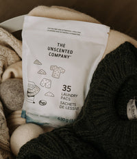 A white bag of 35 laundry pacs by "The Unscented Company" sitting in a laundry basket with knit sweaters and wool dryer balls.