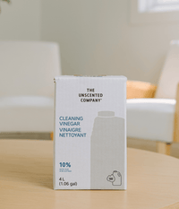 Une boîte blanche de recharge de 4 litres (1,06 gal) de vinaigre nettoyant à 10 % de la marque The Unscented Company, posée sur une table en bois clair dans un salon au décor épuré et lumineux.