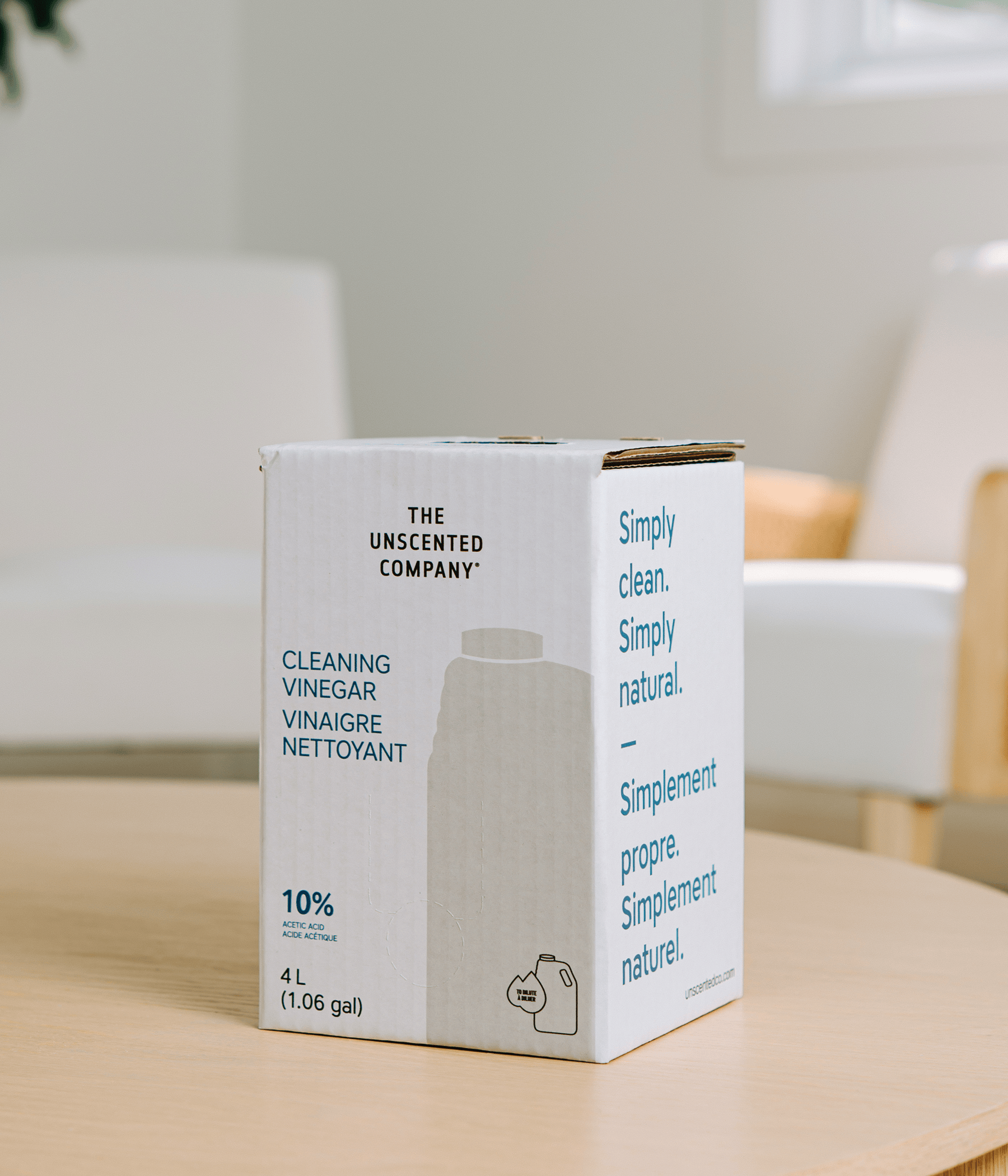 Une boîte blanche de recharge de 4 litres (1,06 gal) de vinaigre nettoyant à 10 % de la marque The Unscented Company, posée sur une table en bois clair dans un salon au décor épuré et lumineux.
