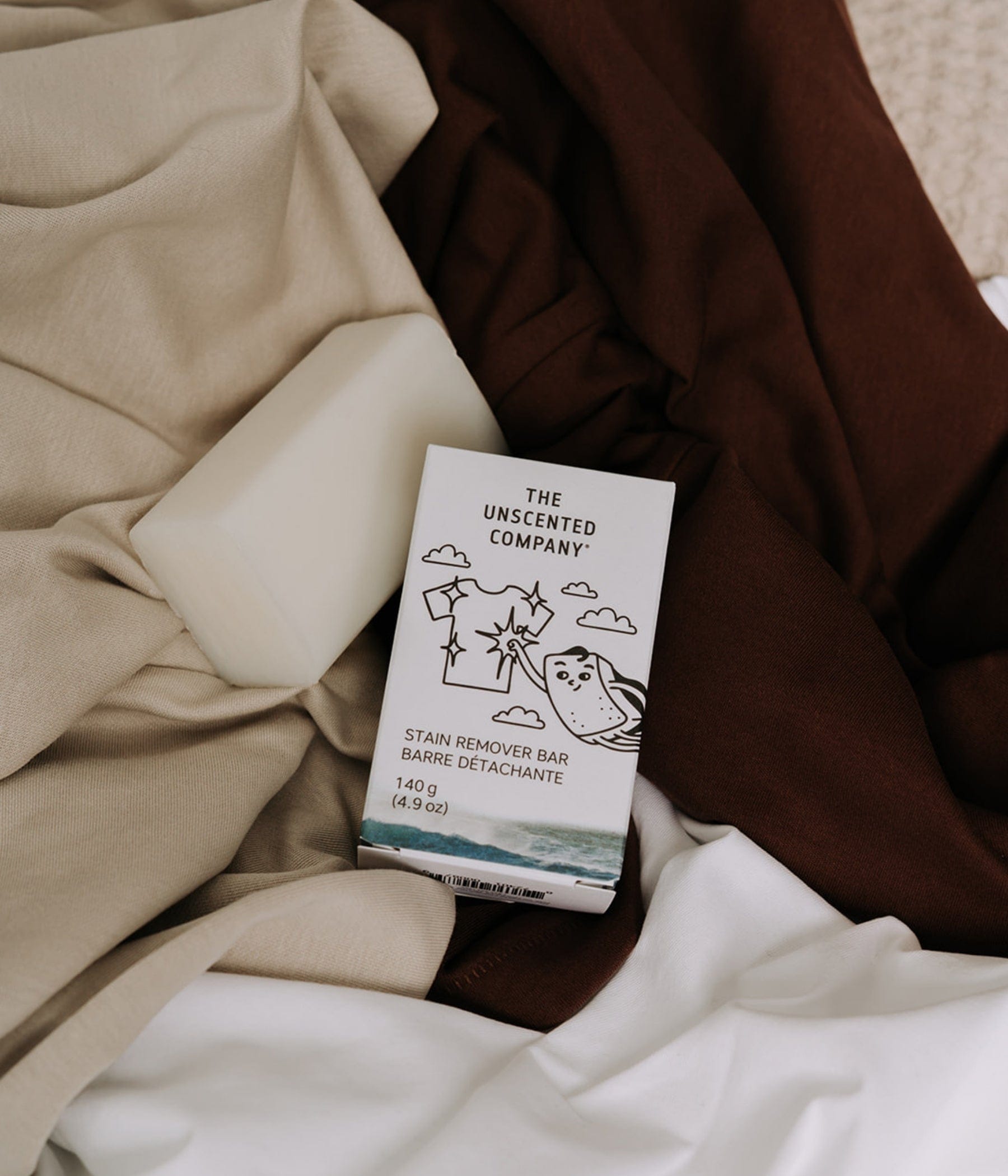 An unwrapped white stain remover bar and its The Unscented Company box (140 g) resting on an arrangement of folded brown, beige, and white fabrics.