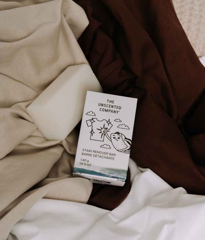 An unwrapped white stain remover bar and its The Unscented Company box (140 g) resting on an arrangement of folded brown, beige, and white fabrics.