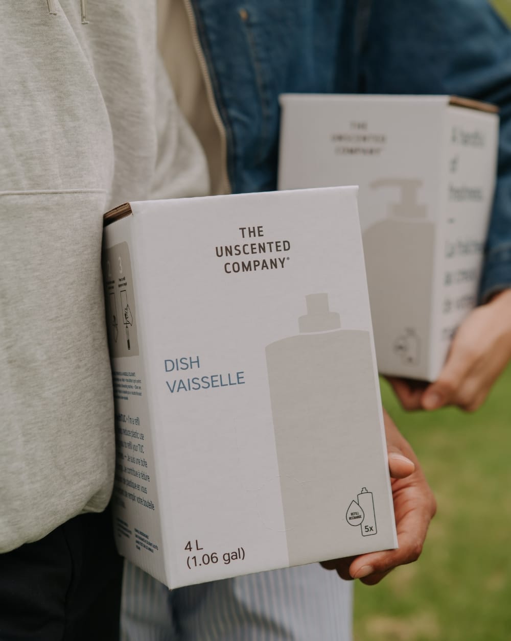 Two people each holding a The Unscented Company 4L refill box — Dish Soap in the foreground — outdoors on a grassy background.