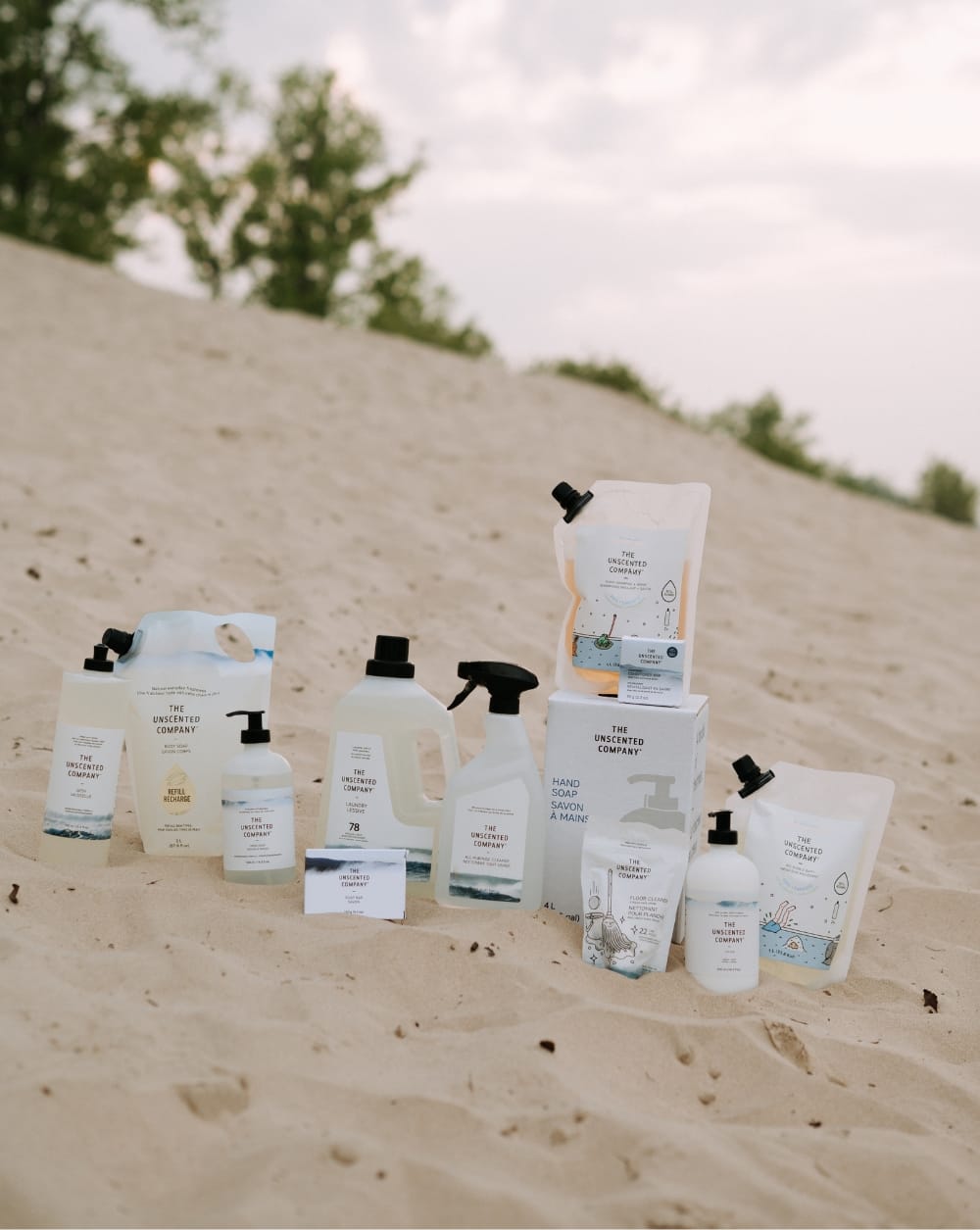 A wide range of The Unscented Company products displayed on a sandy dune, including body soap, laundry detergent, all-purpose cleaner, hand soap, floor cleaner, soap bars, and various refill pouches and bottles.
