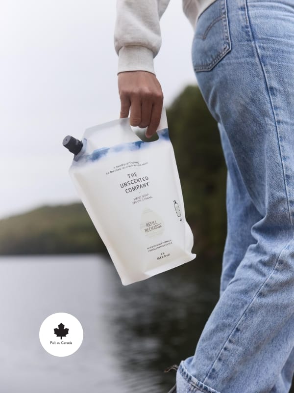 Hand holding a refill pouch of The Unscented Company hand soap labeled “Refill Recharge,” with “Fait au Canada” badge in the corner. Background shows water and blurred trees.