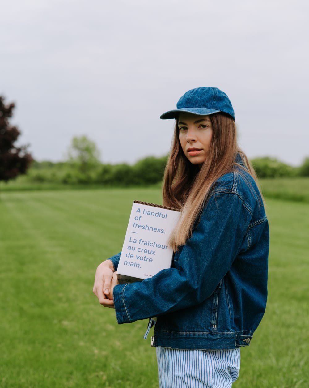 A young woman in a denim jacket and blue cap standing in a green field, holding a The Unscented Company refill pouch with the bilingual tagline "A handful of freshness. — La fraîcheur au creux de votre main."