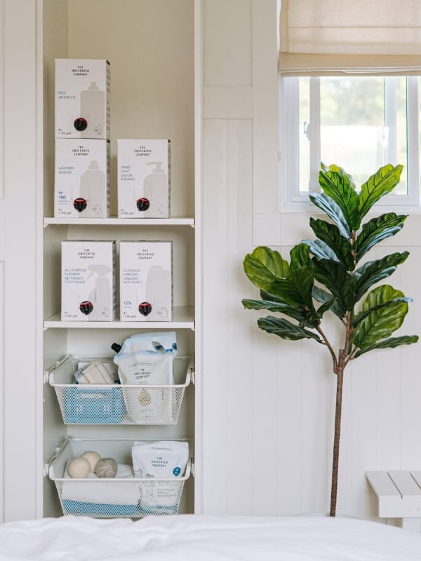 A well-organized white laundry room shelf storing The Unscented Company refill boxes — including dish soap, laundry detergent, hand soap, all-purpose cleaner, and cleaning vinegar — alongside wire baskets holding smaller products and dryer balls, next to a fiddle-leaf fig plant by a window.